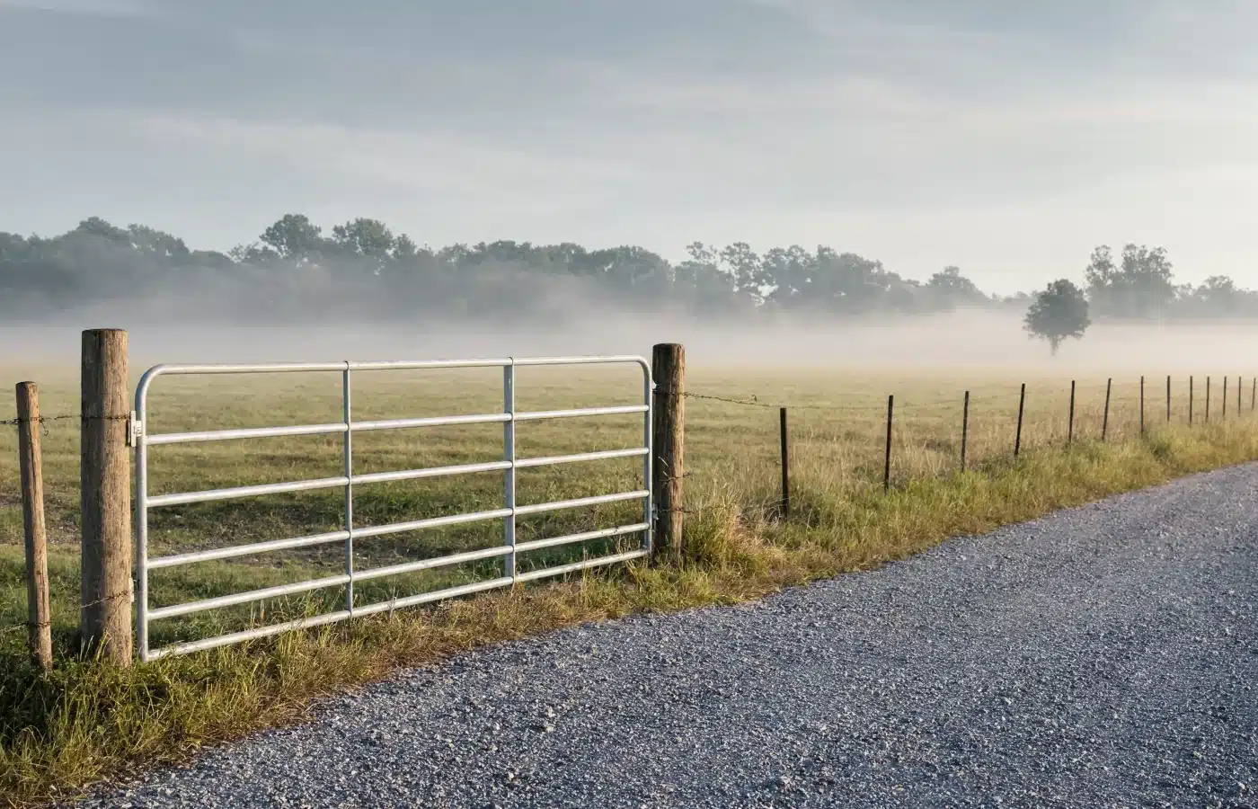A closed galvanized steel farm gate on a gravel pull-off, with a barbed-wire fence line running into an open field