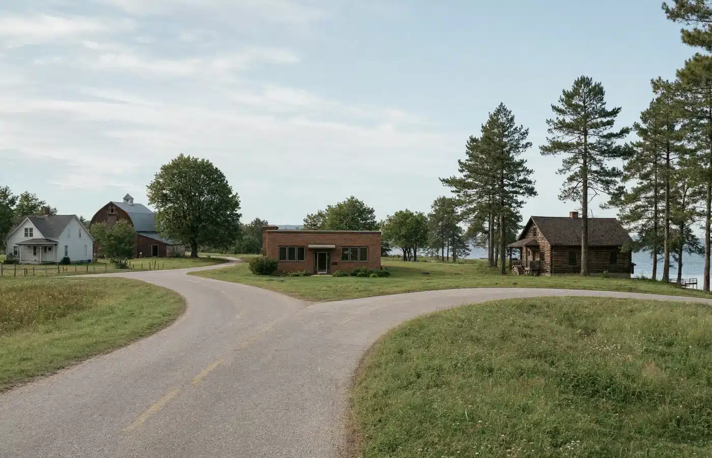 A rural Wisconsin crossroads with a farmhouse and barn, a small brick commercial building and a log cabin by the lake