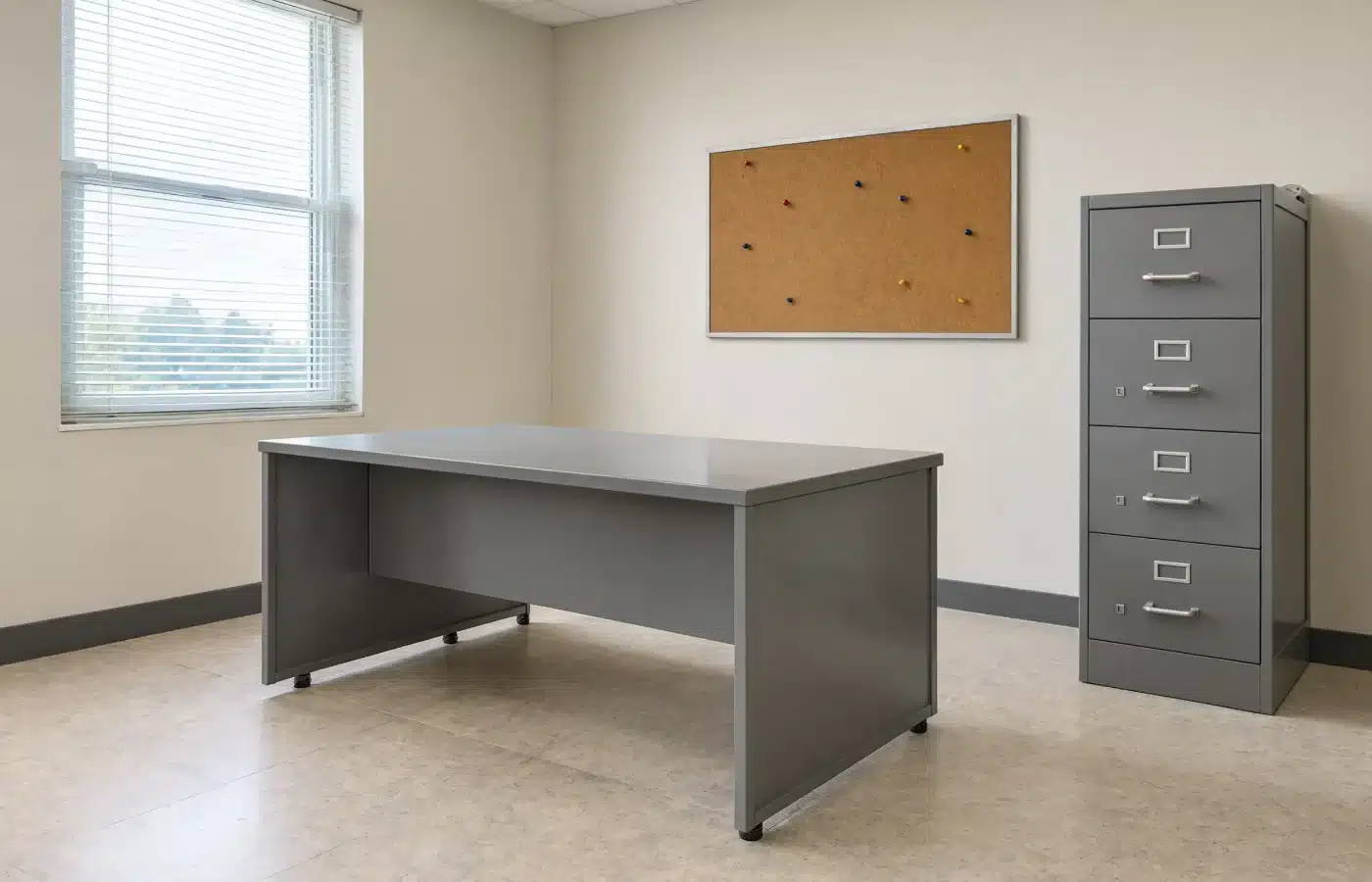 An empty back office with a metal desk, a four-drawer file cabinet and a cork board left on the wall