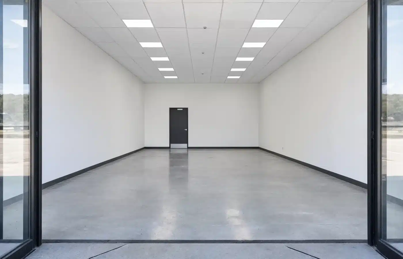 An empty retail bay seen through the storefront glass, with a polished concrete floor and a back service door