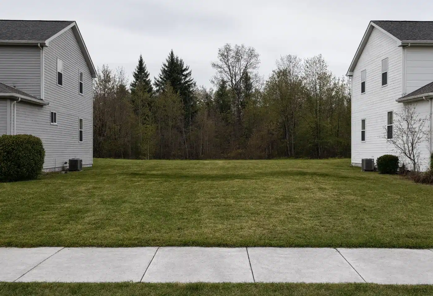A narrow mown vacant lot wedged between two suburban houses with no driveway
